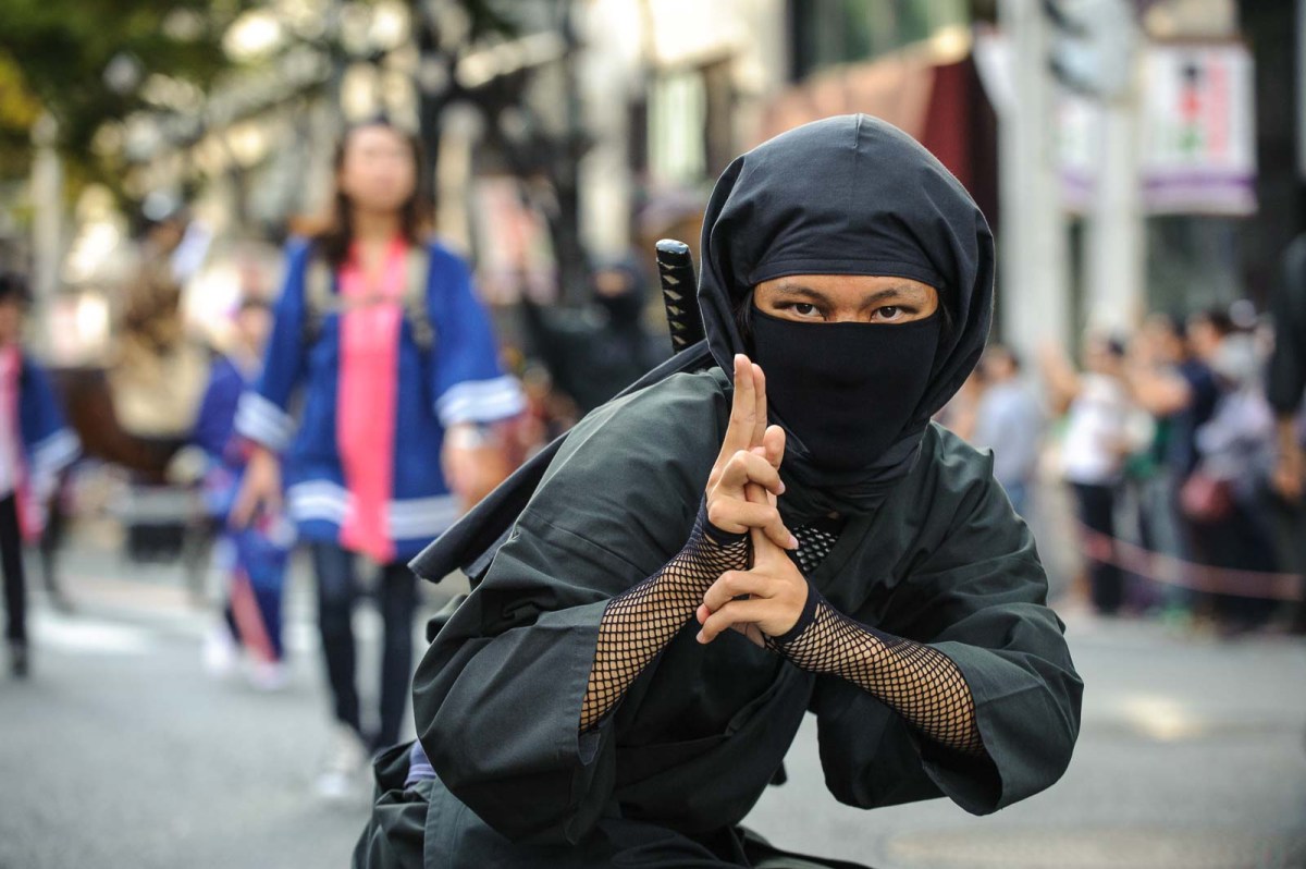 A man dressed in a ninja suit looks directly at the photographer during the Nagoya Festival parade.